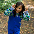 A child smiles, wearing Originals Waterproof Dungarees Blue with a patterned jumper. The child has their hands up and is outside in a garden.